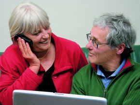 Egy laptop előtt ülő, idős pár egymásra néz, miközben a nő telefonon beszél